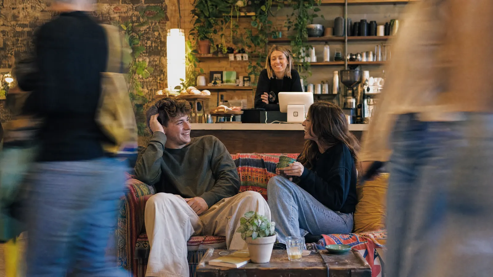 Two people relaxing on a couch in a plant-filled café while the owner works behind the counter.