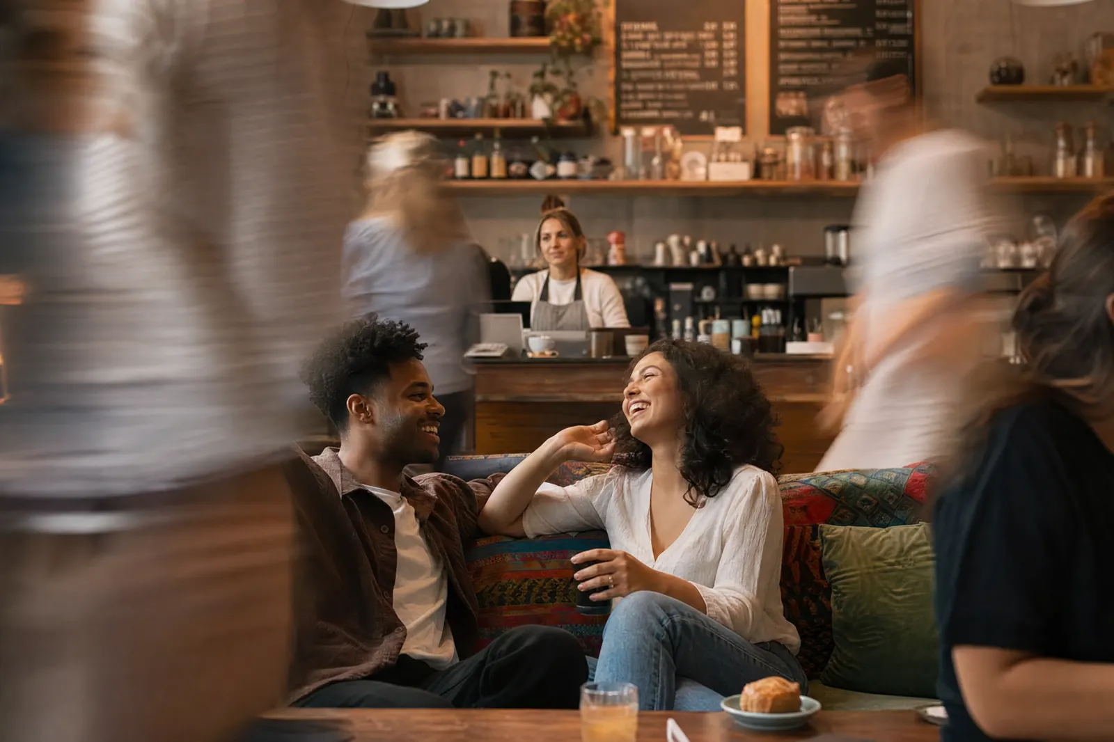 Two friends laughing on a café sofa while people move past in the foreground.