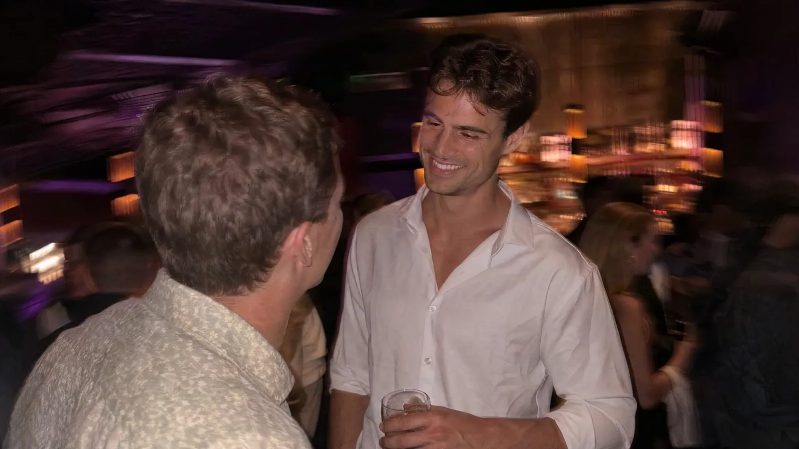 Two friends laughing at a busy bar with warm lights behind them
