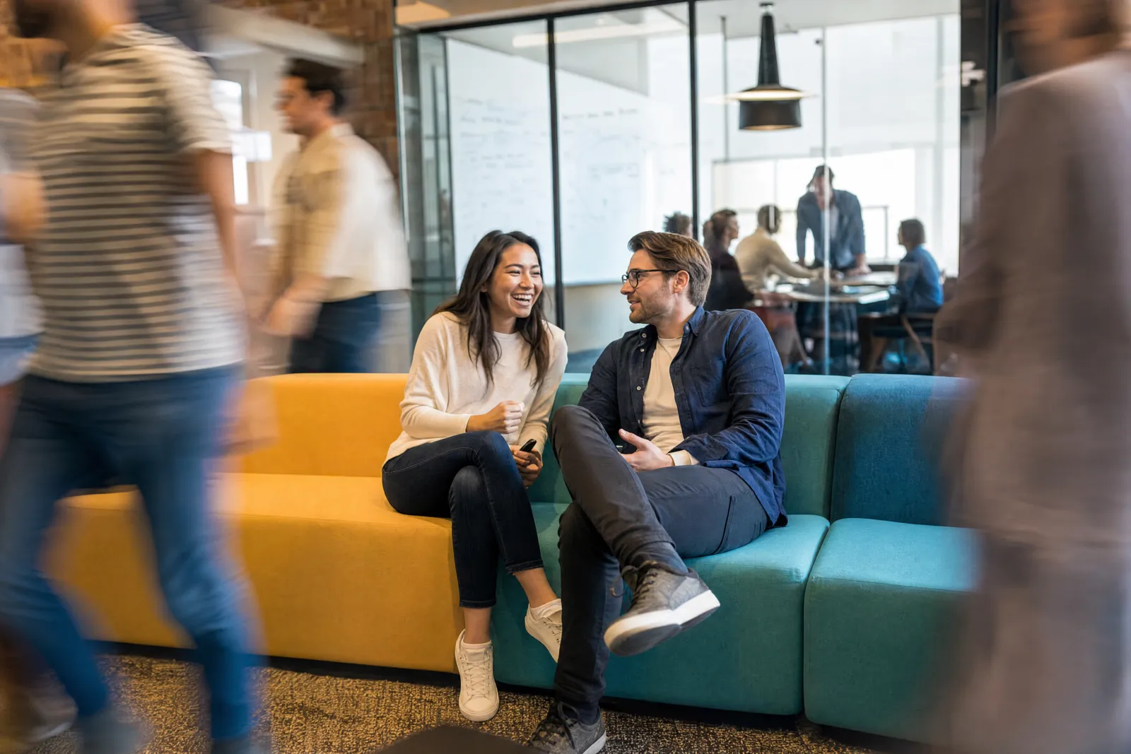 Two colleagues talking on a couch in a busy open office