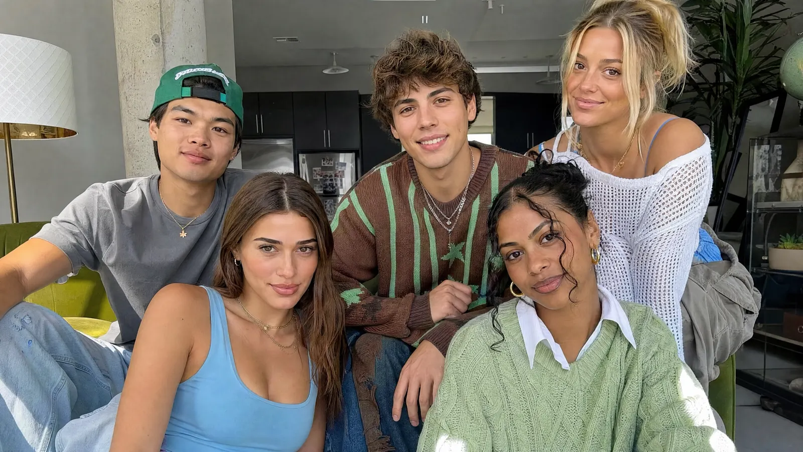 Five friends leaning into frame on a green sofa, one in a backwards cap, all looking at the camera.