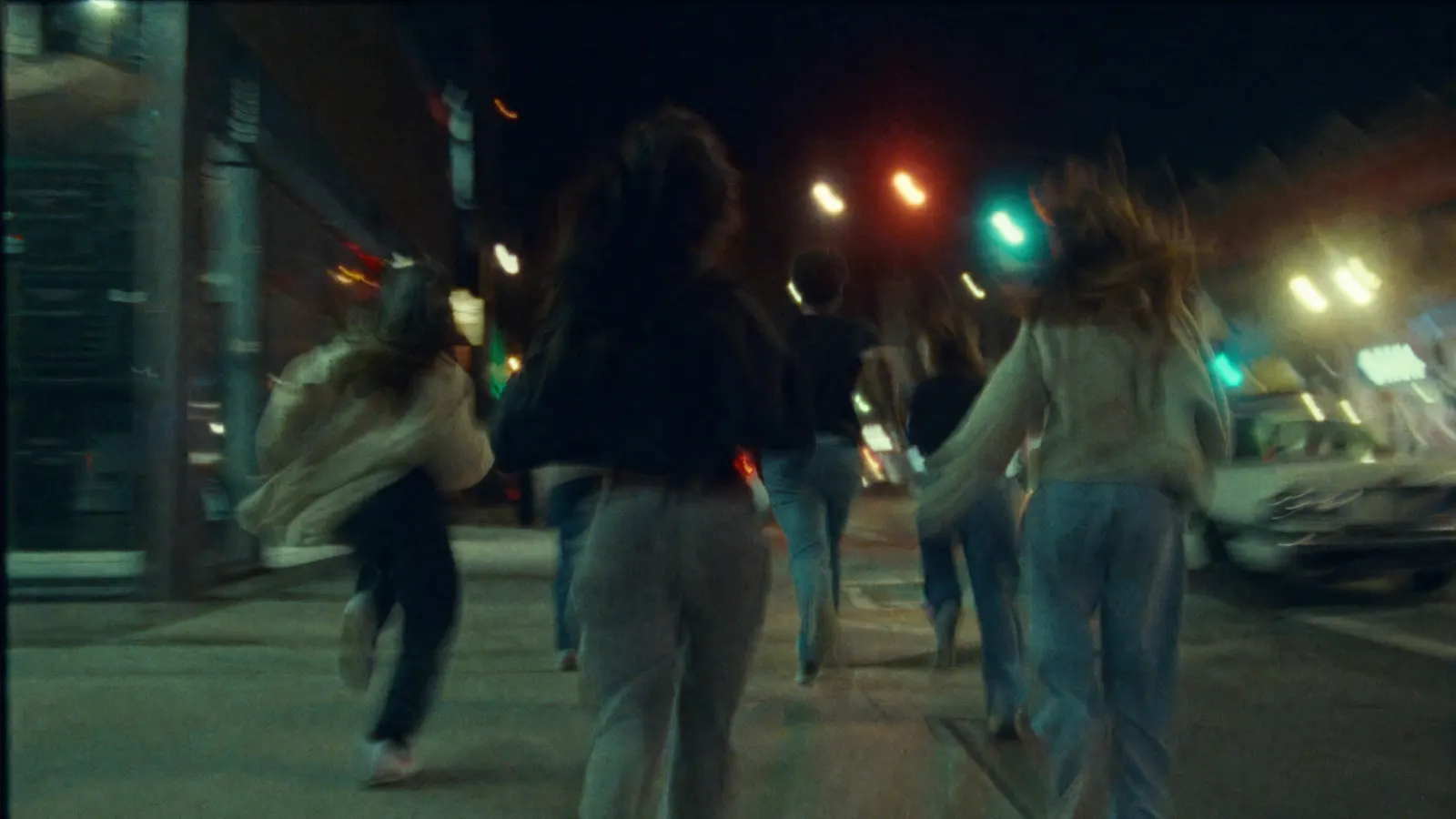 A group of friends running down a city street at night under traffic lights and storefront glow.